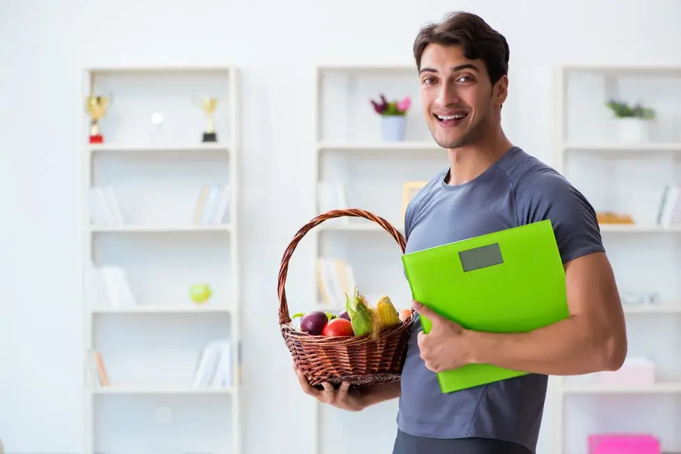 Smiling man holding a basket of vegetables and a green folder, promoting the benefits of eating vegetables as part of a healthy lifestyle at Gym Plus.